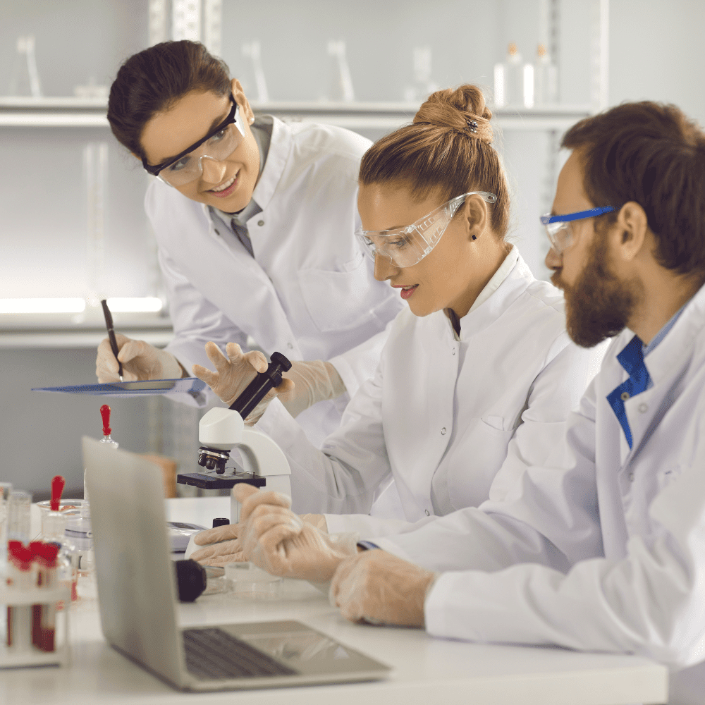 Three people in lab coats and goggles working on a laptop.