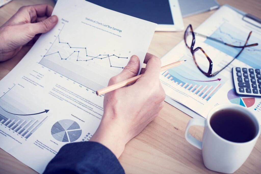 Person analyzing financial charts and graphs on a desk with a pencil in hand, next to a cup of coffee and eyeglasses.