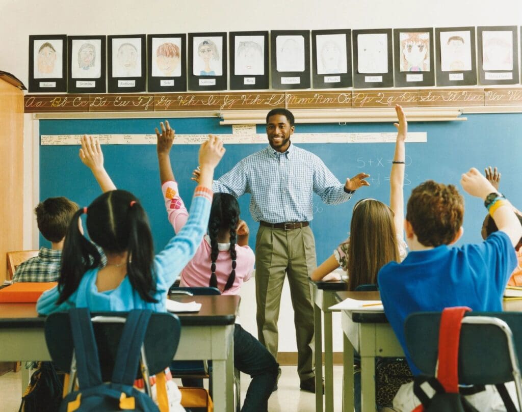 A teacher stands smiling at the front of a classroom while diverse students raise their hands eagerly.