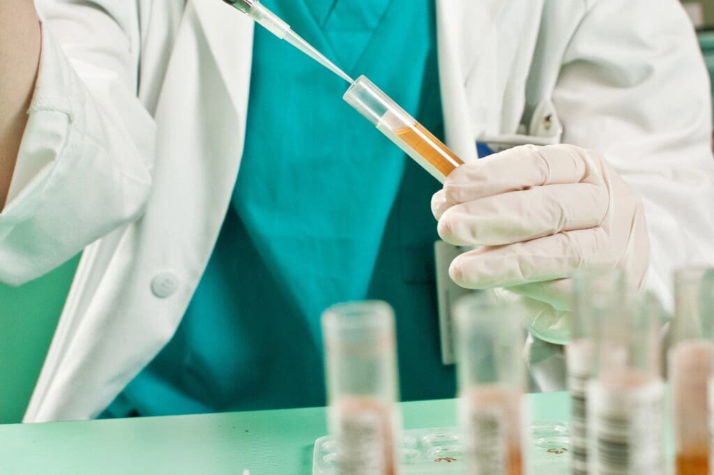 A scientist in a lab coat using a pipette to transfer a liquid into a test tube, surrounded by other lab equipment.