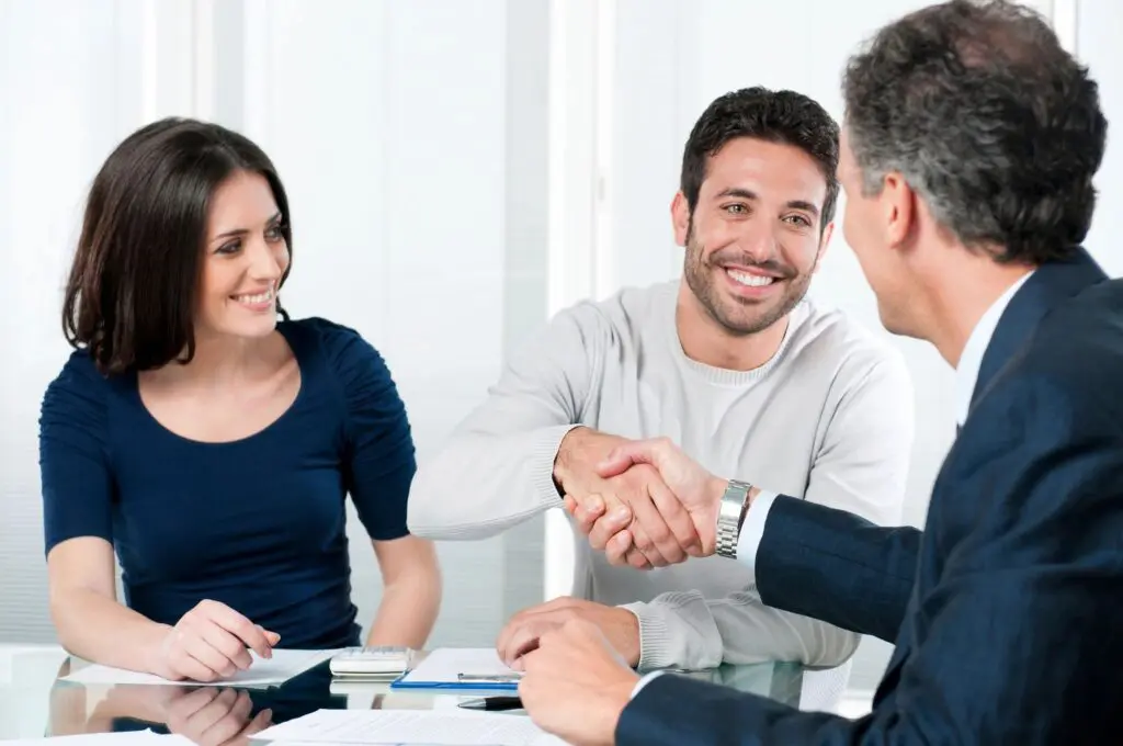 Two men shaking hands at a meeting with a woman smiling at them; documents and a digital tablet on the table.