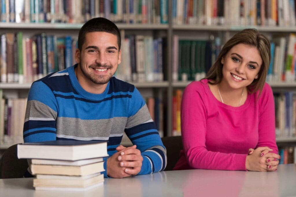 Two students smiling at the camera while sitting at a table with books in a library.