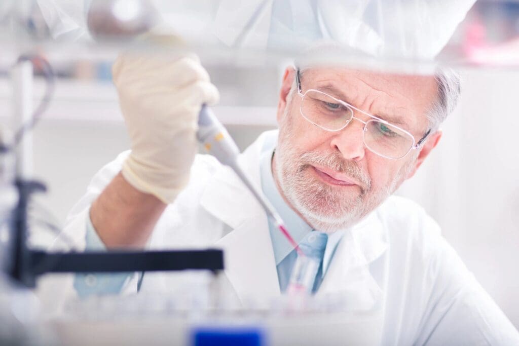 Senior male scientist in lab coat and safety glasses using a pipette in a laboratory setting.