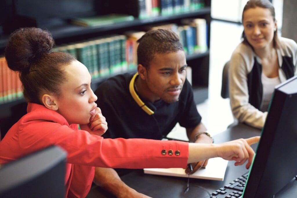 Three young adults collaborate at a computer in a library, focusing intently on the screen.