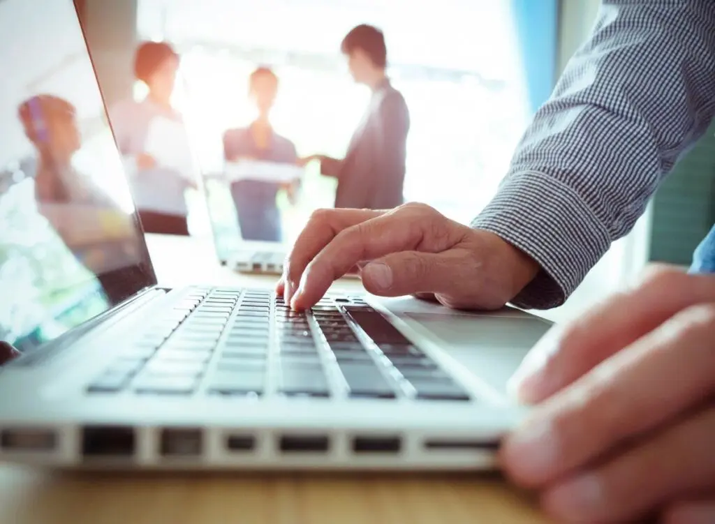 Close-up of a person's hands typing on a laptop in a brightly lit office meeting with colleagues in the background.