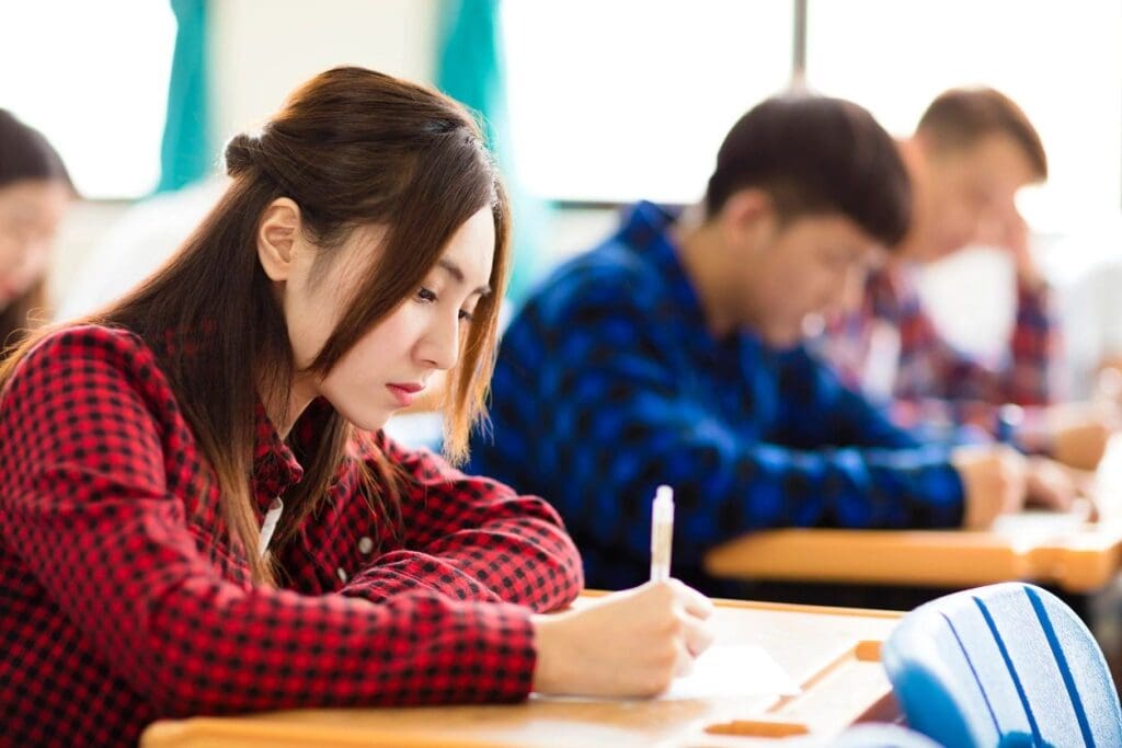 A young asian woman in a red plaid shirt focused on writing notes in a classroom with other students studying in the background.