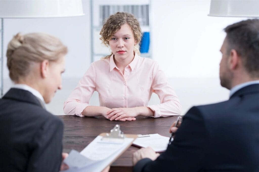 Young woman in a pink shirt sits at a table during a job interview with two interviewers, one female and one male, in a modern office.