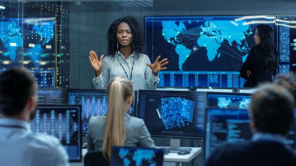 A black woman presenting in a high-tech control room with world maps and data screens, addressing colleagues seated in front of her.