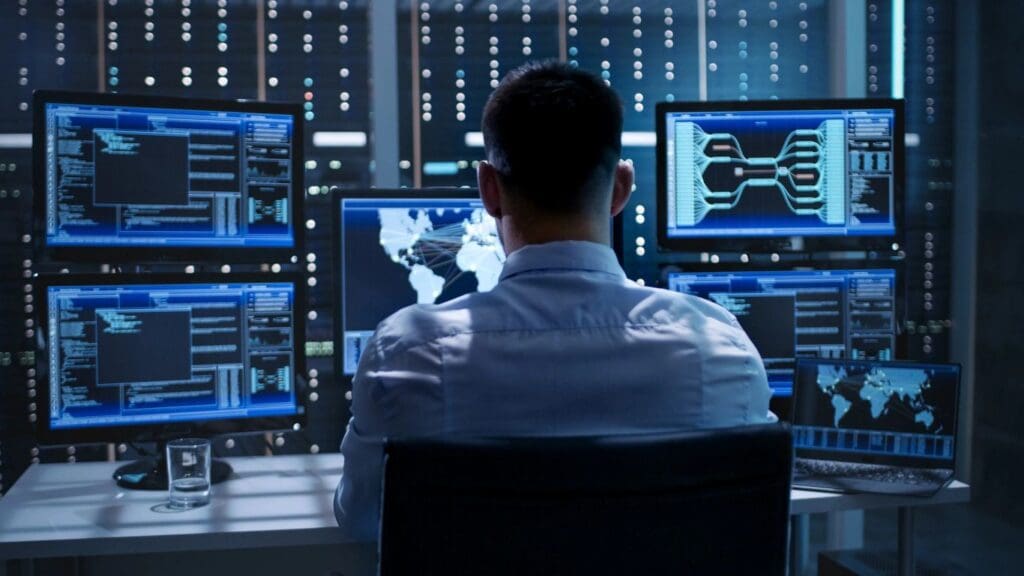 A man sitting in front of multiple computer screens displaying data and graphics in a high-tech control room.