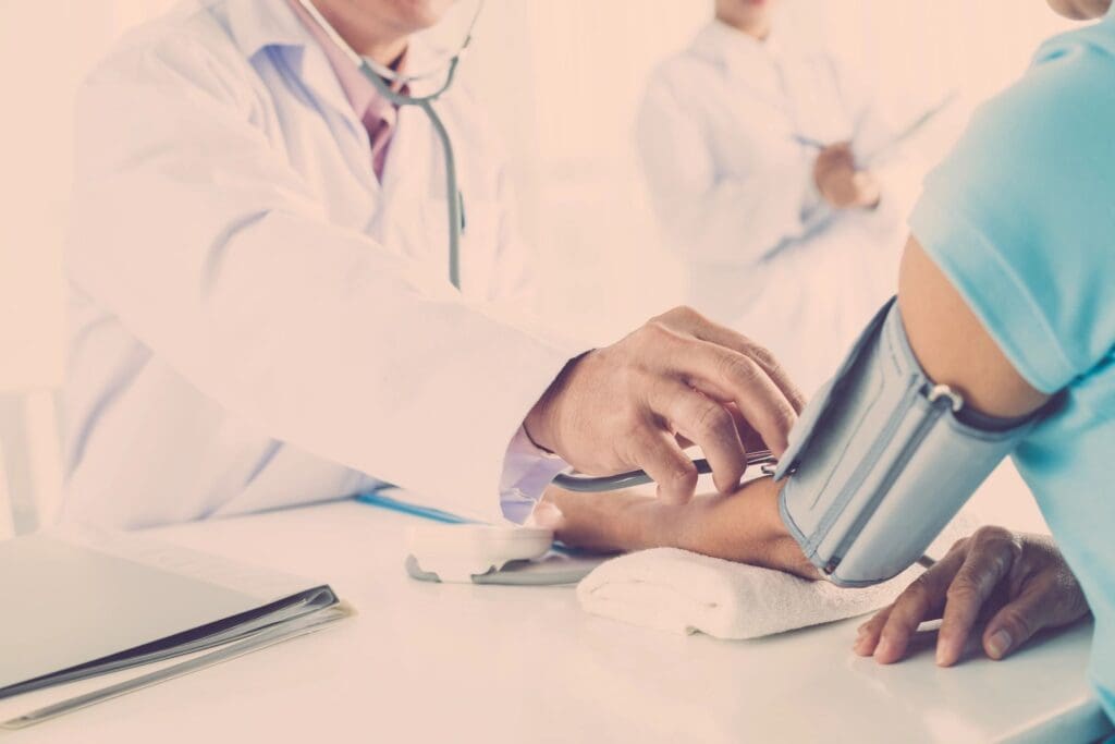 A doctor in a white coat checking a patient's blood pressure with a sphygmomanometer in a clinic, with another healthcare professional discussing online education in the background.