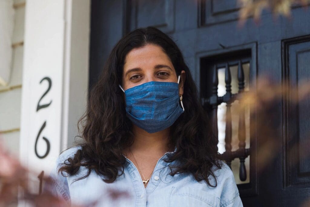 Woman wearing a blue mask and denim shirt stands in front of a house numbered 26, partially obscured by foliage, holding a tablet featuring an online education course.