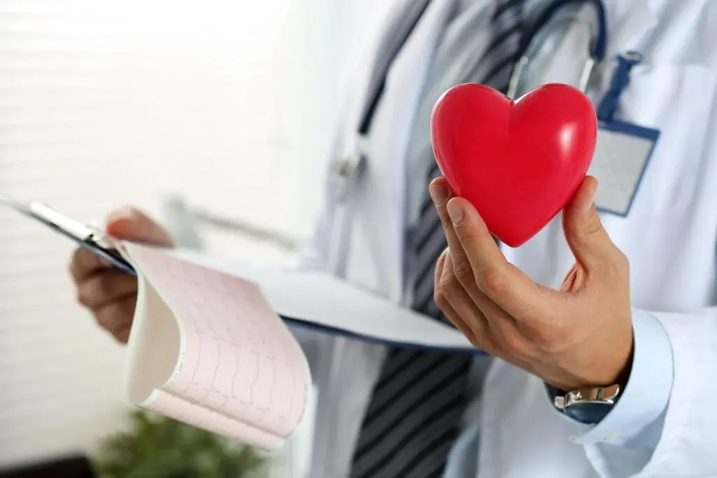 A doctor in a white coat holding a red heart model in one hand and an electrocardiogram strip in the other, demonstrating online education methods.