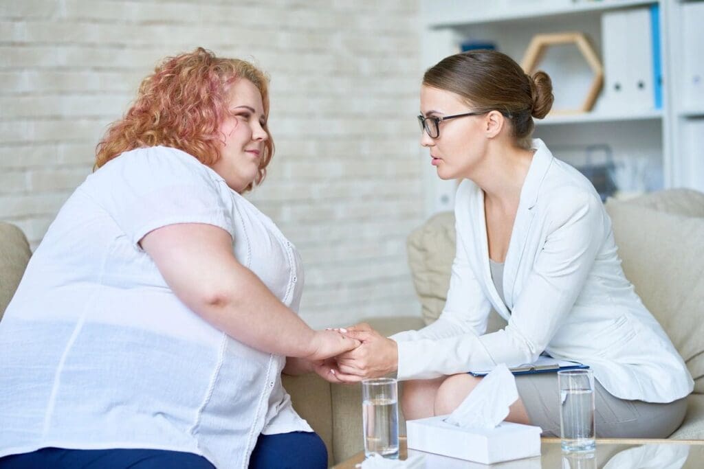 Two women having a conversation on a couch about online education, one in a white suit holding the hands of the other, who is wearing a white blouse.