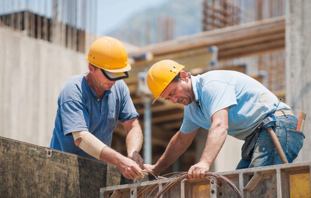 Two construction workers in hard hats and blue shirts are bending and tying rebar at a construction site.