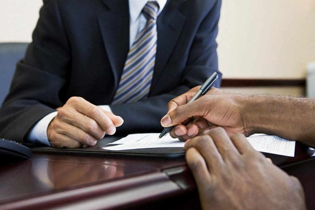 Two individuals in business attire seated at a desk, reviewing and signing documents. only their hands and torsos are visible.