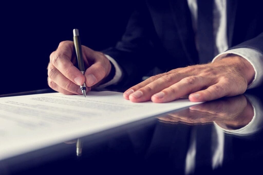 A man in a suit signing a document on a dark table, focusing on his hands and the pen.