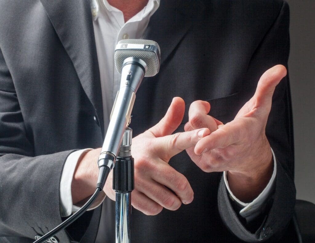 A man in a suit gesturing with his hand while speaking into a microphone.