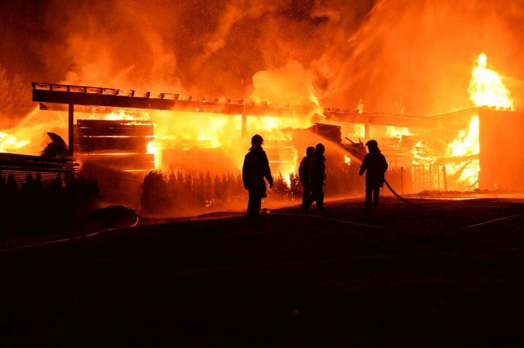 Firefighters standing in front of a massive blaze at night, actively working to extinguish a large building fire.