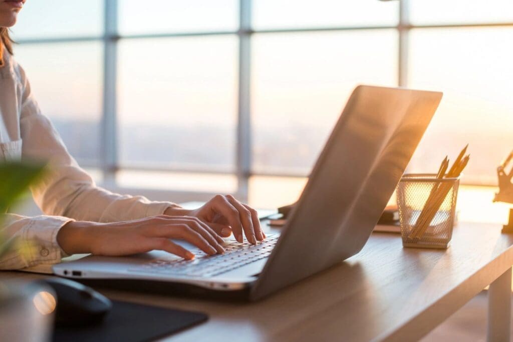 Woman typing on a laptop at a desk during sunset, with a window and office supplies in the background.