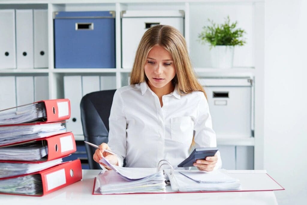 A focused woman in a white shirt examines documents at her office desk, using a calculator and surrounded by binders.