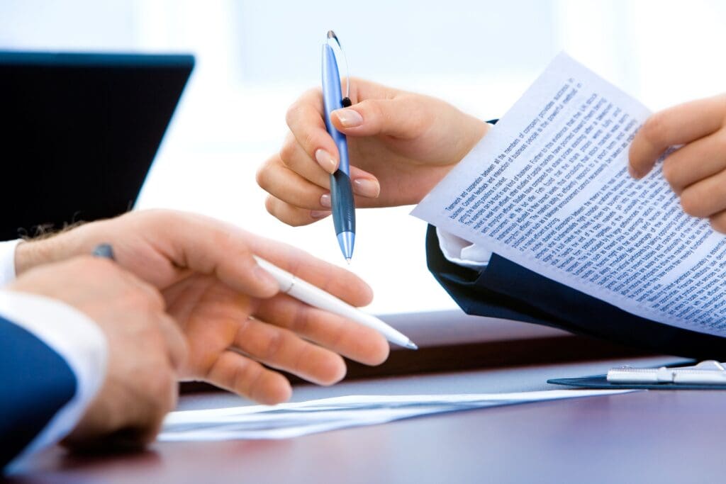 Two business professionals discussing a document at a desk, one handing over a pen for the other to sign.