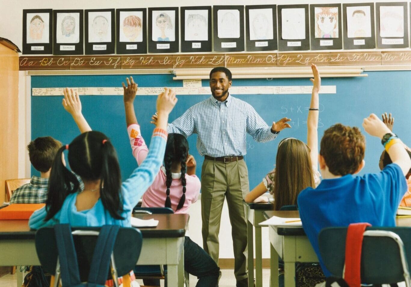 A teacher stands smiling at the front of a classroom while diverse students raise their hands eagerly.
