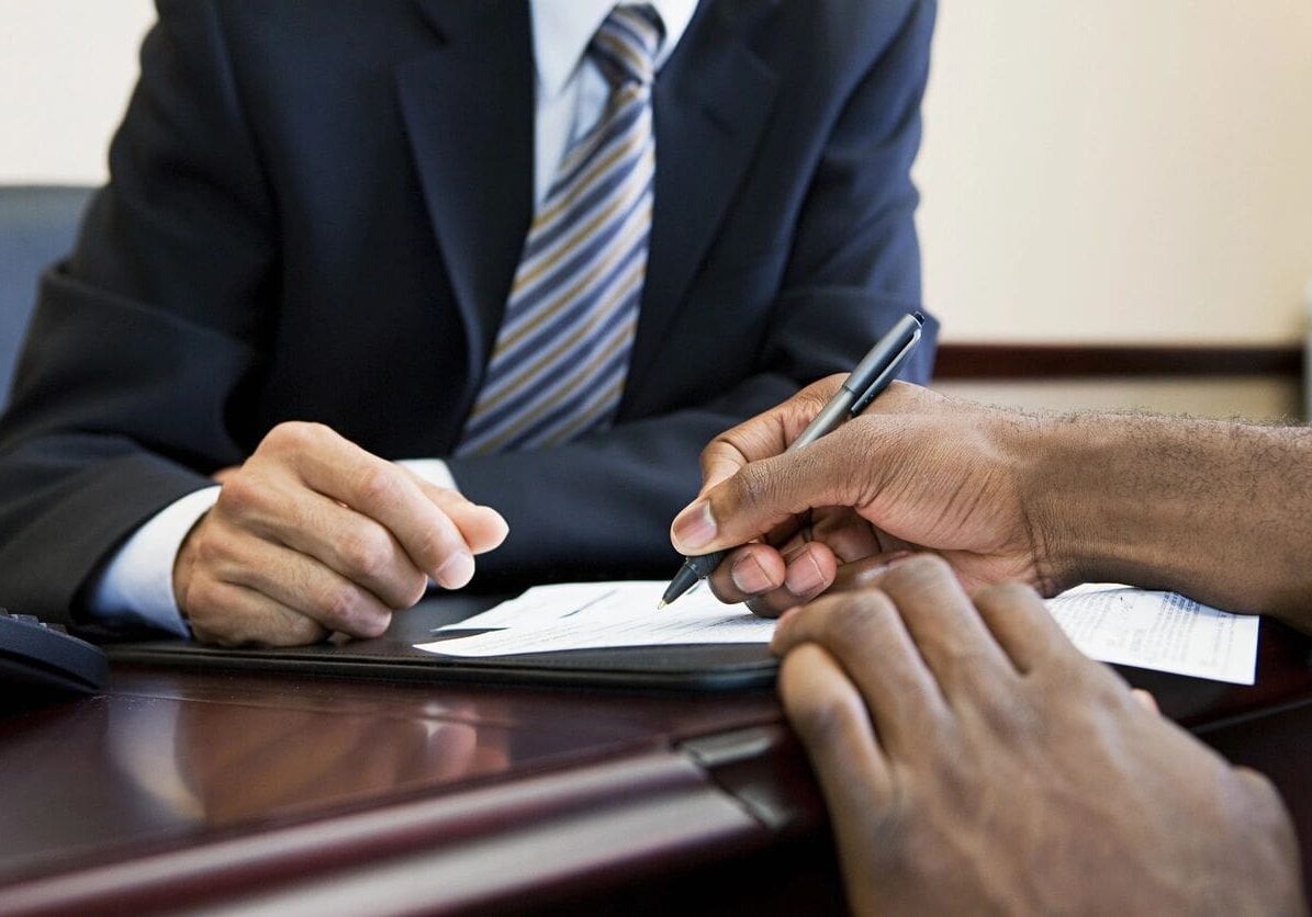 Two individuals in business attire seated at a desk, reviewing and signing documents. only their hands and torsos are visible.