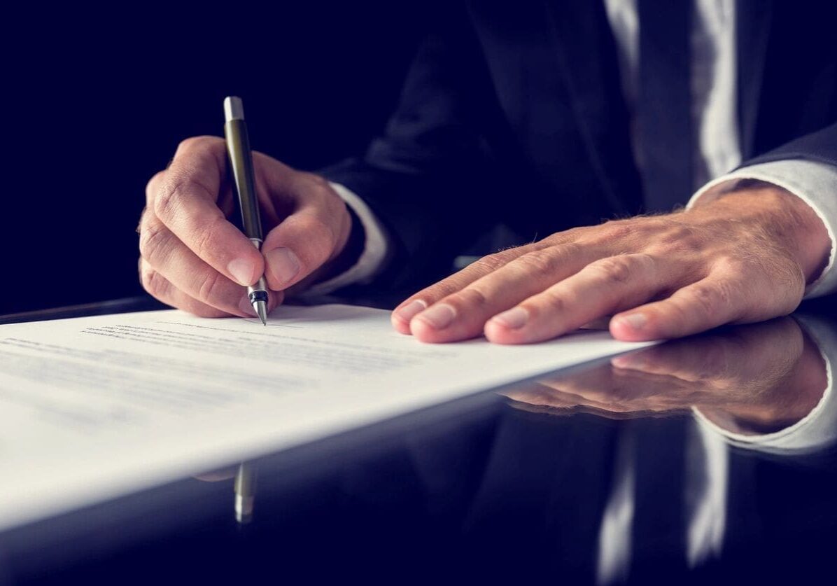 A man in a suit signing a document on a dark table, focusing on his hands and the pen.