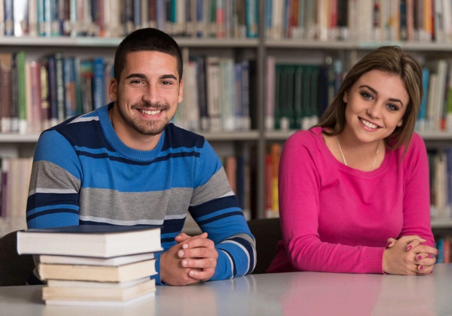 Two students smiling at the camera while sitting at a table with books in a library.