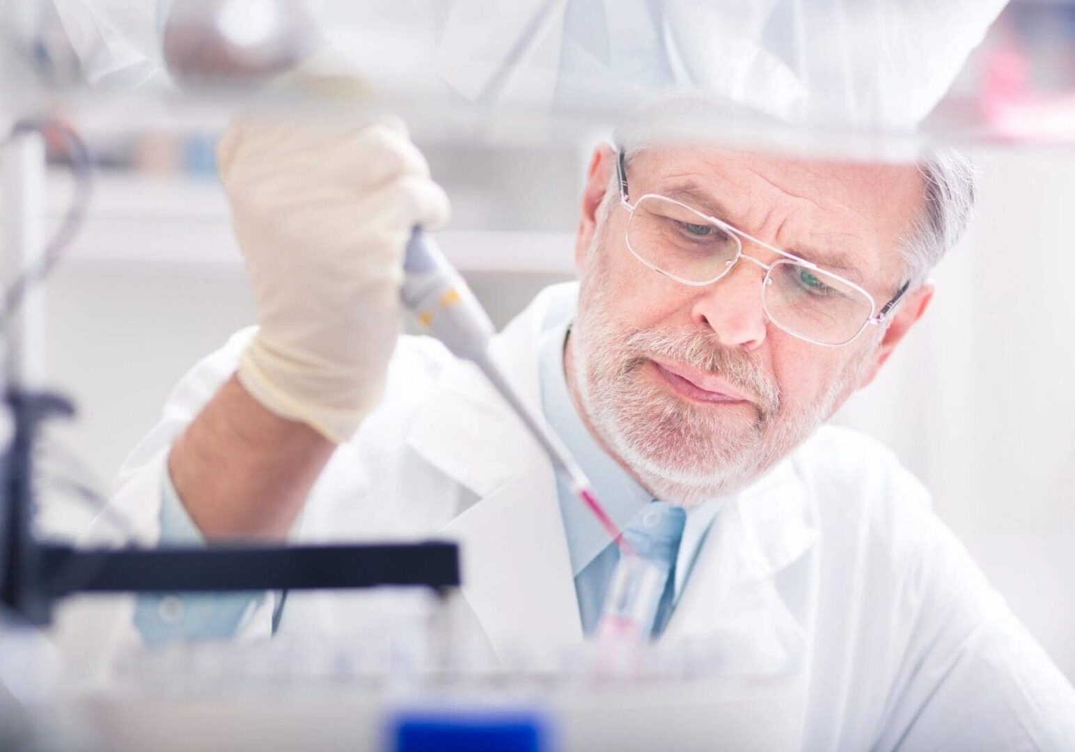 Senior male scientist in lab coat and safety glasses using a pipette in a laboratory setting.