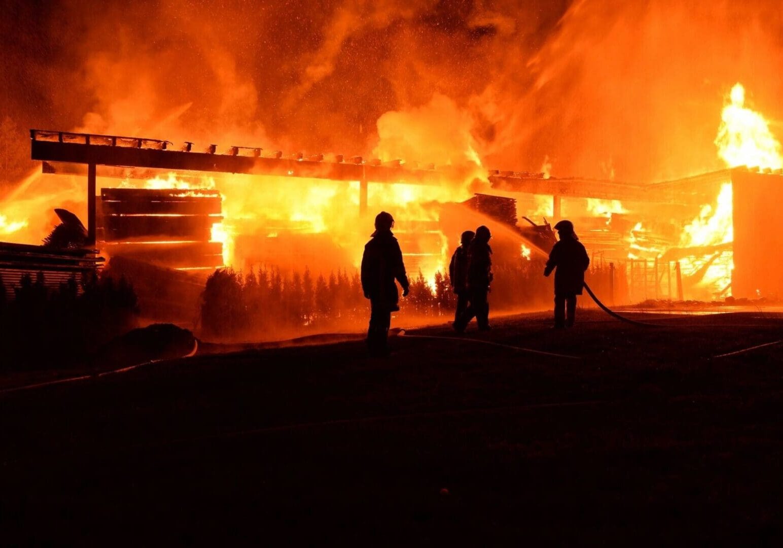 Firefighters standing in front of a massive blaze at night, actively working to extinguish a large building fire.