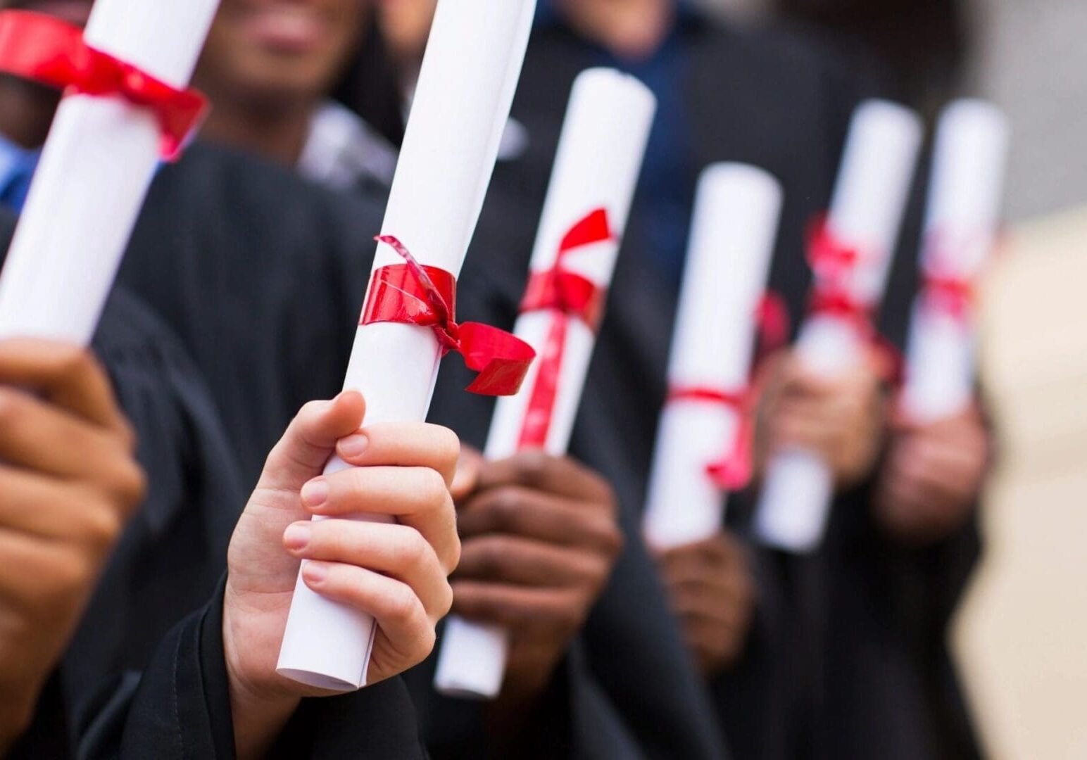 Graduates in black robes holding diplomas with red ribbons, close-up of their hands and diplomas.