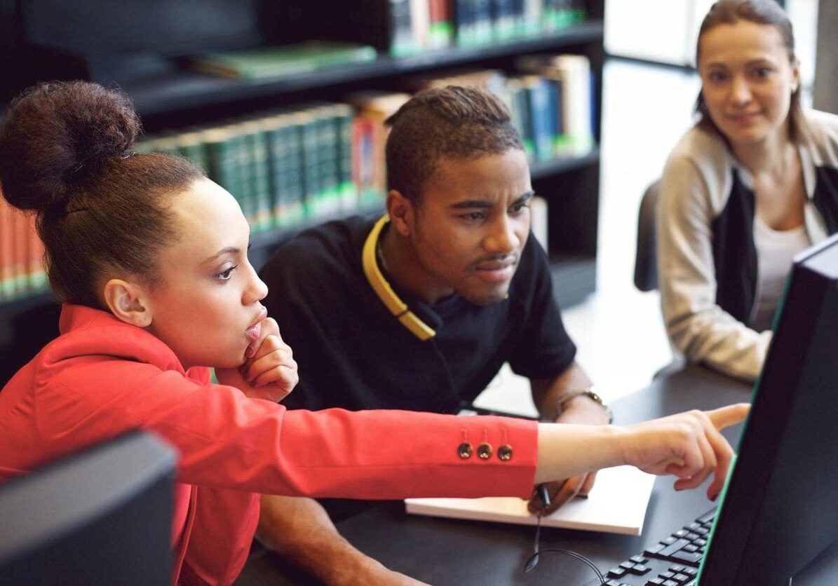 Three young adults collaborate at a computer in a library, focusing intently on the screen.