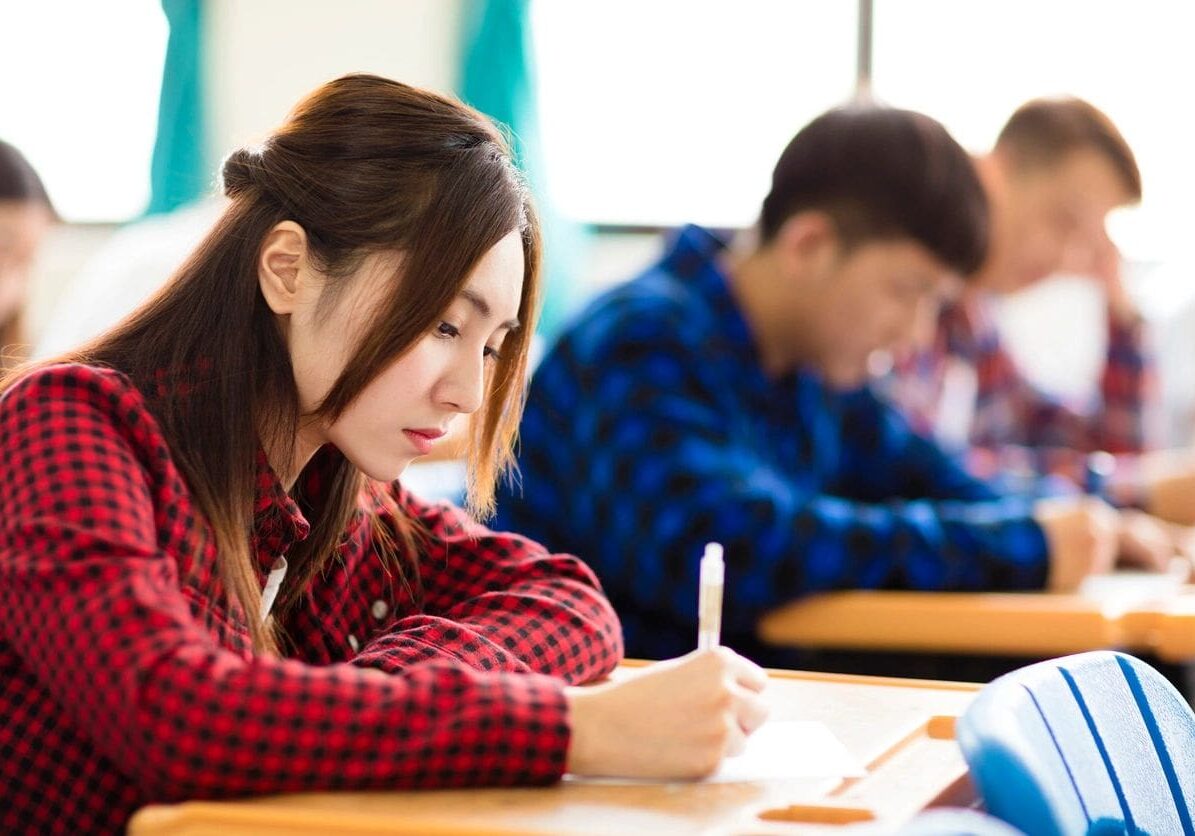 A young asian woman in a red plaid shirt focused on writing notes in a classroom with other students studying in the background.