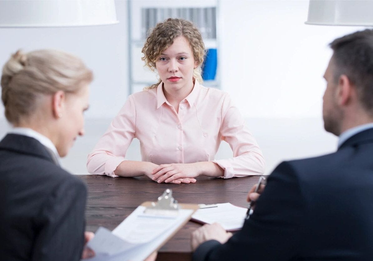Young woman in a pink shirt sits at a table during a job interview with two interviewers, one female and one male, in a modern office.