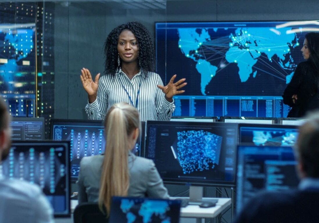 A black woman presenting in a high-tech control room with world maps and data screens, addressing colleagues seated in front of her.