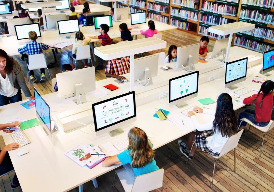 Modern library setting with students of diverse ages using computers and reading, supervised by an adult.