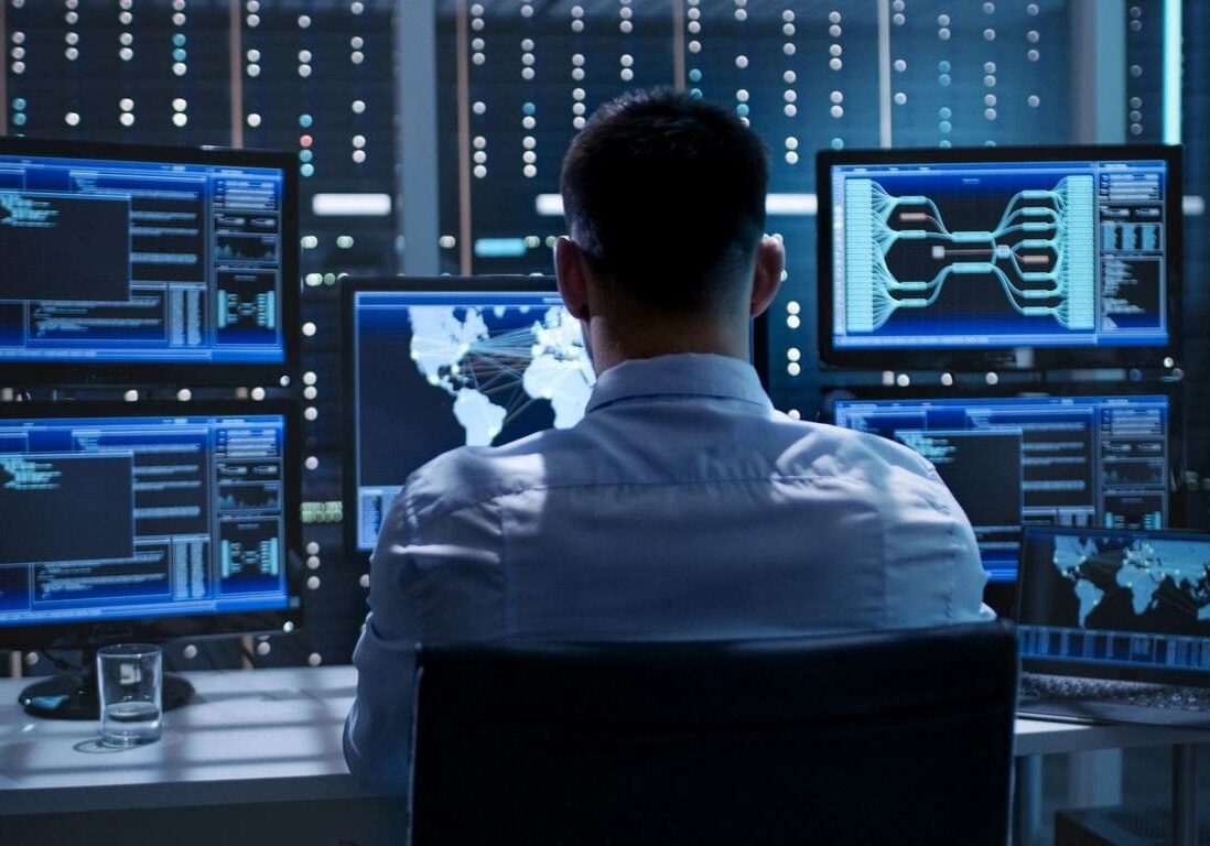 A man sitting in front of multiple computer screens displaying data and graphics in a high-tech control room.