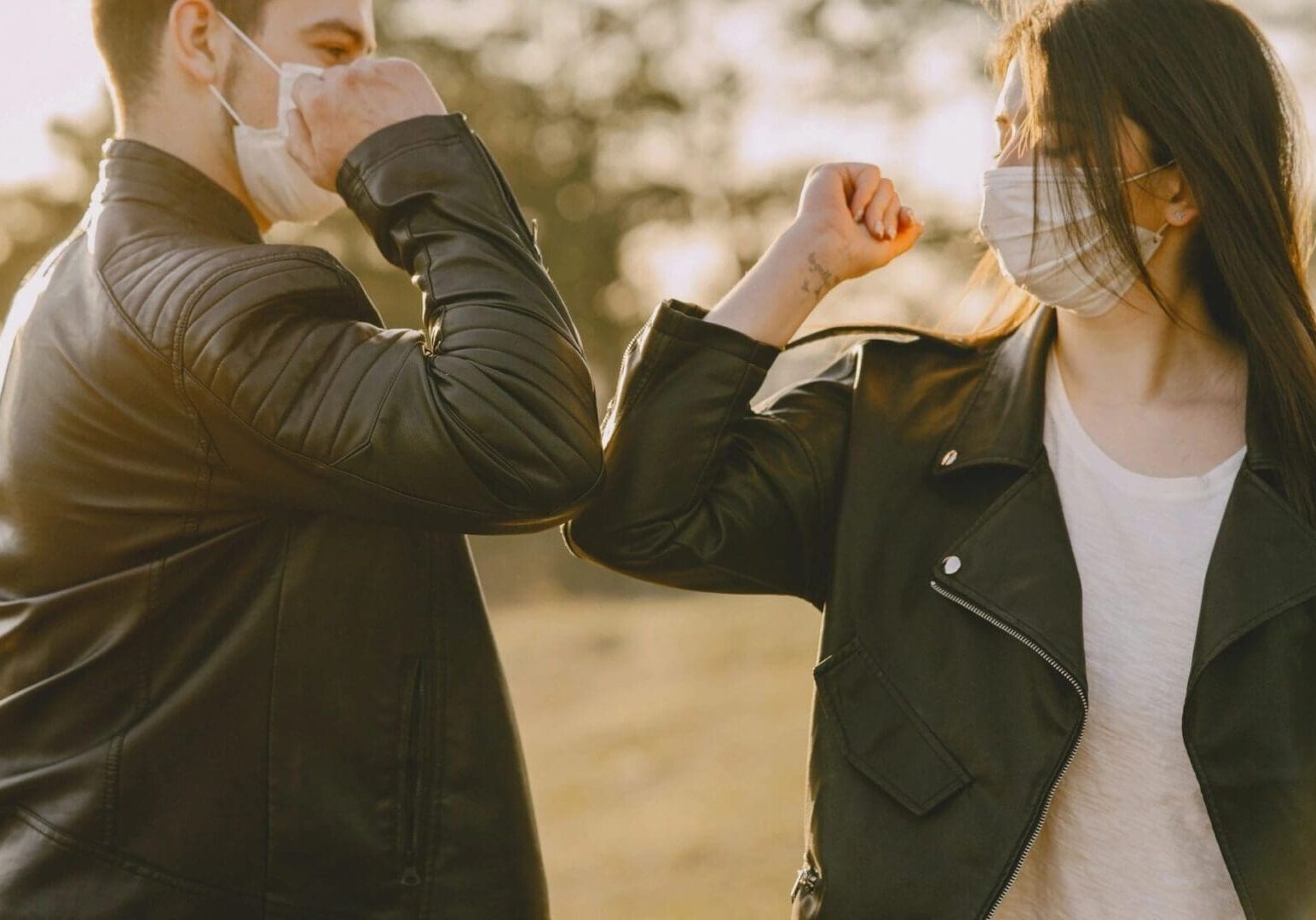 Two people in leather jackets and face masks greeting each other with an elbow bump outdoors at sunset after an online education session.